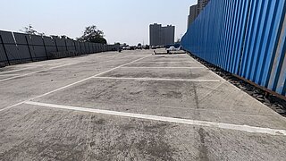Leerer Parkplatz mit weißen Markierungen auf Betonboden zwischen einem blauen Zaun rechts und einem grauen Zaun links, im Hintergrund Hochhäuser und Bäume