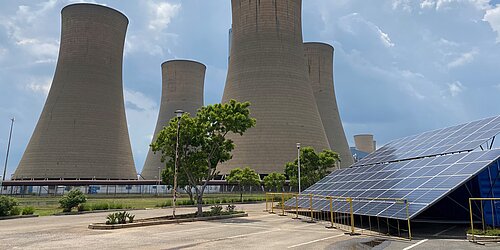 Ein großes Kohlekraftwerk mit vier monumentalen Kühltürmen dominiert das Bild. Im Vordergrund befinden sich Solarzellen auf einer schrägen Konstruktion, umgeben von einem Zaun. Einige Bäume und Büsche sorgen für etwas Grün in der Szene. Der Himmel ist leicht bewölkt.