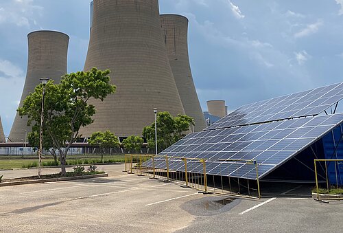 A large coal power plant with four monumental cooling towers dominates the image. In the foreground, solar panels are mounted on a tilted structure, enclosed by a fence. Some trees and shrubs add greenery to the scene. The sky is partly cloudy.