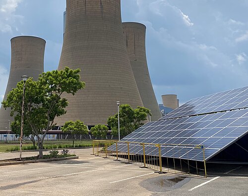 A large coal power plant with four monumental cooling towers dominates the image. In the foreground, solar panels are mounted on a tilted structure, enclosed by a fence. Some trees and shrubs add greenery to the scene. The sky is partly cloudy.