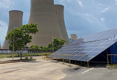 Ein großes Kohlekraftwerk mit vier monumentalen Kühltürmen dominiert das Bild. Im Vordergrund befinden sich Solarzellen auf einer schrägen Konstruktion, umgeben von einem Zaun. Einige Bäume und Büsche sorgen für etwas Grün in der Szene. Der Himmel ist leicht bewölkt.