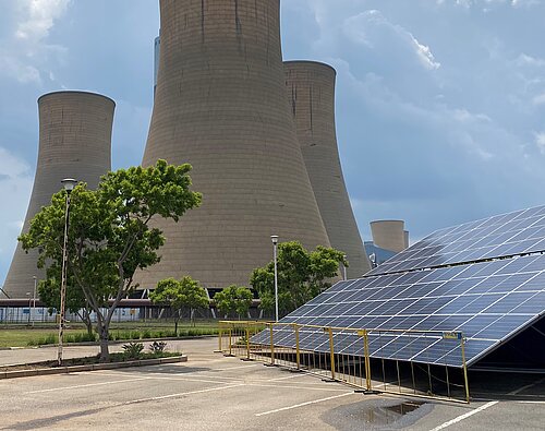 Ein großes Kohlekraftwerk mit vier monumentalen Kühltürmen dominiert das Bild. Im Vordergrund befinden sich Solarzellen auf einer schrägen Konstruktion, umgeben von einem Zaun. Einige Bäume und Büsche sorgen für etwas Grün in der Szene. Der Himmel ist leicht bewölkt.