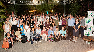 Ein Gruppenfoto zeigt alle Teilnehmenden des IKI‑Netzwerkabends. Alle tragen Namensschilder um den Hals. Am Bildrand sind grüne Pflanzen zu sehen und daneben stehen Würfel mit dem IKI‑Logo.