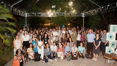 Ein Gruppenfoto zeigt alle Teilnehmenden des IKI‑Netzwerkabends. Alle tragen Namensschilder um den Hals. Am Bildrand sind grüne Pflanzen zu sehen und daneben stehen Würfel mit dem IKI‑Logo.