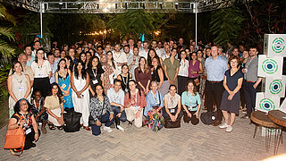 A group photo shows all participants of the IKI network evening. Everyone wears name tags around the neck. Green plants are visible at the edge of the image and beside them are cubes bearing the IKI logo.