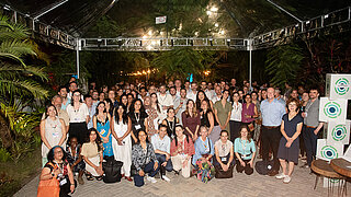 A group photo shows all participants of the IKI network evening. Everyone wears name tags around the neck. Green plants are visible at the edge of the image and beside them are cubes bearing the IKI logo.