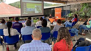 Several people sit under a tent watching a presentation about the Nicoya Peninsula projected on a screen. A woman is giving the talk at a table with an orange tablecloth.