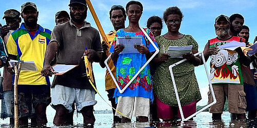 A group of people stands in the shallow ocean waters of Papua New Guinea, taking part in a training session to monitor seagrass meadows and mangrove forests.