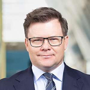 Ein Mann mit kurzem Haar und Brille steht in einem modernen Innenraum. Er trägt einen dunkelblauen Anzug, ein weißes Hemd und eine gestreifte Krawatte. Seine Hände sind vor dem Körper verschränkt. Im Hintergrund sind unscharfe, farbige Wandflächen und eine große, helle Wand sichtbar.