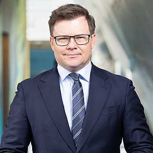 A man with short hair and glasses stands in a modern indoor setting. He is wearing a dark blue suit, a white shirt, and a striped tie. His hands are clasped in front of his body. The background features blurred, colorful wall surfaces and a large, light wall.