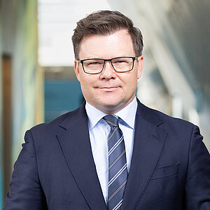 Ein Mann mit kurzem Haar und Brille steht in einem modernen Innenraum. Er trägt einen dunkelblauen Anzug, ein weißes Hemd und eine gestreifte Krawatte. Seine Hände sind vor dem Körper verschränkt. Im Hintergrund sind unscharfe, farbige Wandflächen und eine große, helle Wand sichtbar.