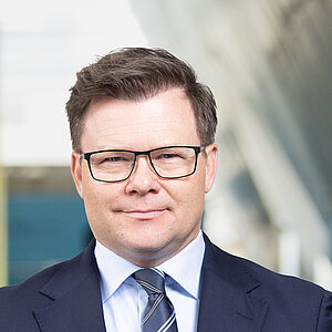 A man with short hair and glasses stands in a modern indoor setting. He is wearing a dark blue suit, a white shirt, and a striped tie. His hands are clasped in front of his body. The background features blurred, colorful wall surfaces and a large, light wall.