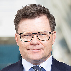 A man with short hair and glasses stands in a modern indoor setting. He is wearing a dark blue suit, a white shirt, and a striped tie. His hands are clasped in front of his body. The background features blurred, colorful wall surfaces and a large, light wall.