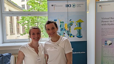 Zwei Frauen lächeln vor Informationsrollups mit Logos der Bundesregierung, IKI, im Hintergrund Fenster mit Blick auf Bäume und Gebäude