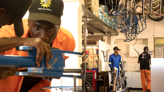 Close-up of a man wearing a cap carefully adjusting a blue bicycle fixture, with a workshop filled with tools, shelves, and bicycles behind him.