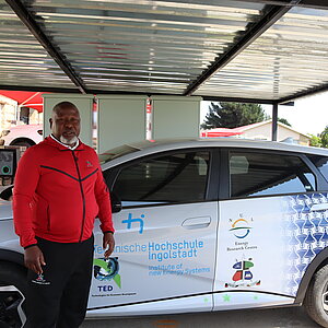 Ein Mann in rotem Oberteil steht vor einem Elektroauto mit Logos der Technischen Hochschule Ingolstadt und des Energy Research Center in Lesotho unter einem Carport.