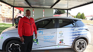 A man wearing a red top stands in front of an electric car with logos of the Technical University of Ingolstadt and the Energy Research Center in Lesotho under a carport.