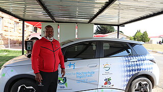 Ein Mann in rotem Oberteil steht vor einem Elektroauto mit Logos der Technischen Hochschule Ingolstadt und des Energy Research Center in Lesotho unter einem Carport.