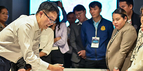 Group of people standing around a table with large sheets of paper and photos, one person pointing at the table.
