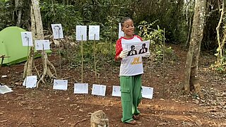Eine Frau mit buntem Oberteil hält ein Blatt mit zwei Porträtzeichnungen vor einer Reihe von Zeichnungen auf Papier im Wald