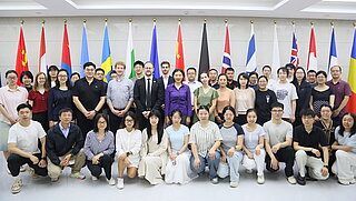 Große Gruppe von Menschen posiert für ein Gruppenfoto in einem hellen Raum, hinter ihnen stehen mehrere internationale Flaggen.