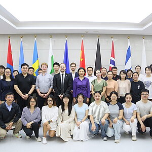 Große Gruppe von Menschen posiert für ein Gruppenfoto in einem hellen Raum, hinter ihnen stehen mehrere internationale Flaggen.