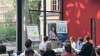 Eine Frau steht an einem Rednerpult und spricht zu einem Publikum von etwa 20 Personen in einem modernen, hellen Konferenzraum mit großen Glasfenstern. Links ist ein Flipchart mit Notizen, rechts ein Roll-up der „International Climate Initiative (IKI)“. Die Zuhörenden sitzen an runden Tischen, auf denen Wasserflaschen, Gläser und Notizmaterialien stehen. Im Hintergrund ist grüne Vegetation vor den Fenstern zu sehen.