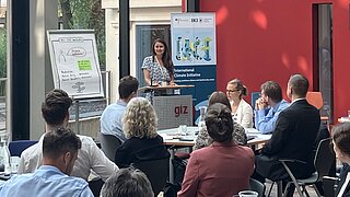 A woman stands at a lectern speaking to an audience of about 20 people in a modern, bright conference room with large glass windows. On the left, there is a flipchart with notes; on the right, a roll-up banner of the "International Climate Initiative (IKI)." The attendees sit at round tables with water bottles, glasses, and notepads. Green vegetation can be seen outside the windows in the background.
