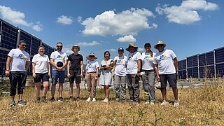 Gruppe von zehn Personen steht auf trockenem Grasfeld zwischen zwei Reihen von Solarpanelen unter blauem Himmel mit wenigen Wolken.