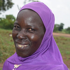 Smiling woman wearing a purple headscarf with blurred greenery in the background