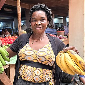 A market vendor is holding a bunch of bananas in each hand and standing in front of a market stall with more bananas and vegetables.