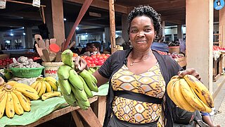 Eine Markhändlerin hält in jeder Hand einen Bund Bananen und steht vor einem Marktstand mit weiteren Bananen und Gemüse.