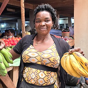 Eine Markhändlerin hält in jeder Hand einen Bund Bananen und steht vor einem Marktstand mit weiteren Bananen und Gemüse.