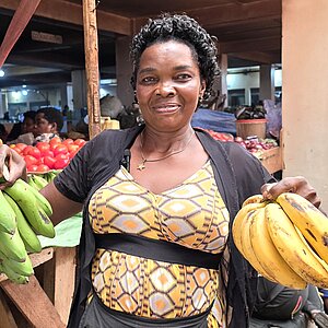 Eine Markhändlerin hält in jeder Hand einen Bund Bananen und steht vor einem Marktstand mit weiteren Bananen und Gemüse.