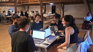 Gruppe junger Menschen sitzt an einem Tisch mit Laptops in einem großen Raum und unterhält sich lachend.