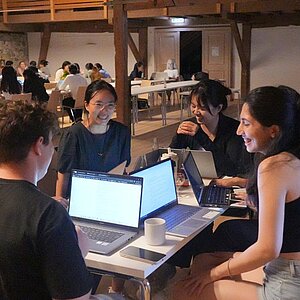 Gruppe junger Menschen sitzt an einem Tisch mit Laptops in einem großen Raum und unterhält sich lachend.