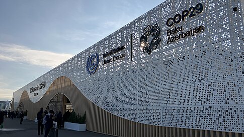  Außenaufnahme des COP29-Geländes in Baku, Aserbaidschan. Das Gebäude hat eine wellenförmige Fassade aus Holz und durchbrochenen, weißen Metallplatten. Logos der Vereinten Nationen, von COP29 und der Regierung Aserbaidschans sind zu sehen. Menschen gehen mit Gepäck und Taschen auf das Gebäude zu.