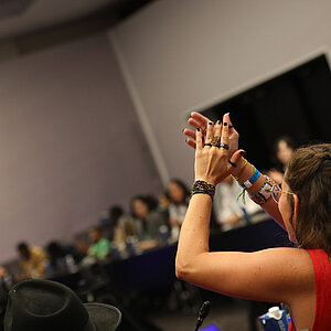 Eine junge Frau steht in einem Konferenzsaal und klatscht über dem Kopf in die Hände.