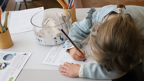 A child with sits at a table, coloring on a piece of paper. On the table are colorful pencils and a jar with paper pouches. In the background, additional papers can be seen, some containing information about the environment and climate protection.