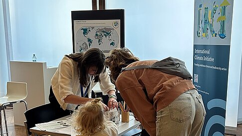 A person is showing a family with a child something on a table. On the table, materials related to environmental and climate protection initiatives are visible, and an informational banner can be seen in the background.