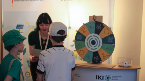 Zwei Kinder stehen vor einem Stand mit einem Glücksrad und einer Frau, die etwas erklärt