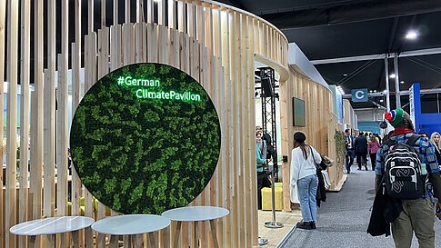 Innenaufnahme des German Climate Pavilion auf der COP29. Im Vordergrund drei runde Stehtische aus Holz. Dahinter eine grüne Mooswand mit der leuchtenden Aufschrift „#German Climate Pavilion“. Die Wände bestehen aus vertikalen Holzlatten. Menschen laufen durch den Gang neben dem Pavillon.