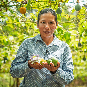 Frau in gepunktetem Hemd hält eine Passionsfrucht und eine Avocado in einem Garten mit vielen Passionsfruchtpflanzen