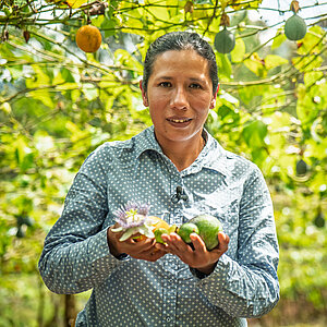 Woman in a patterned shirt holding a passion fruit and an avocado in a garden with many passion fruit plants