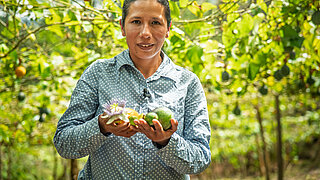 Frau in gepunktetem Hemd hält eine Passionsfrucht und eine Avocado in einem Garten mit vielen Passionsfruchtpflanzen