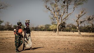 Zwei Frauen in traditioneller Kleidung gehen auf einem trockenen, offenen Feld, eine trägt ein Kind auf dem Rücken, die andere balanciert Gegenstände auf dem Kopf