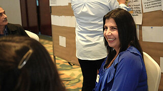 Eine lachende Frau mit dunklen Haaren nimmt an einem Workshop teil, bei dem im Hintergrund Zettel an Pinnwände gehängt werden.