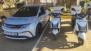 Ein weißes Elektroauto der Marke BYD mit dem Schriftzug 'MaZero' auf der Motorhaube neben zwei weißen Elektrorollern mit 'MaZero'-Schriftzug auf den Fronten, alle auf einem Parkplatz.