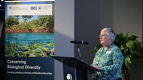 Woman with short gray hair and glasses standing at a podium next to a banner reading 'Conserving Biological Diversity' with an image of water and plants.