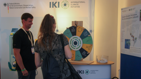 Zwei Personen stehen vor einem Messestand mit einem großen, runden Glücksrad und Informationsplakaten an der Wand